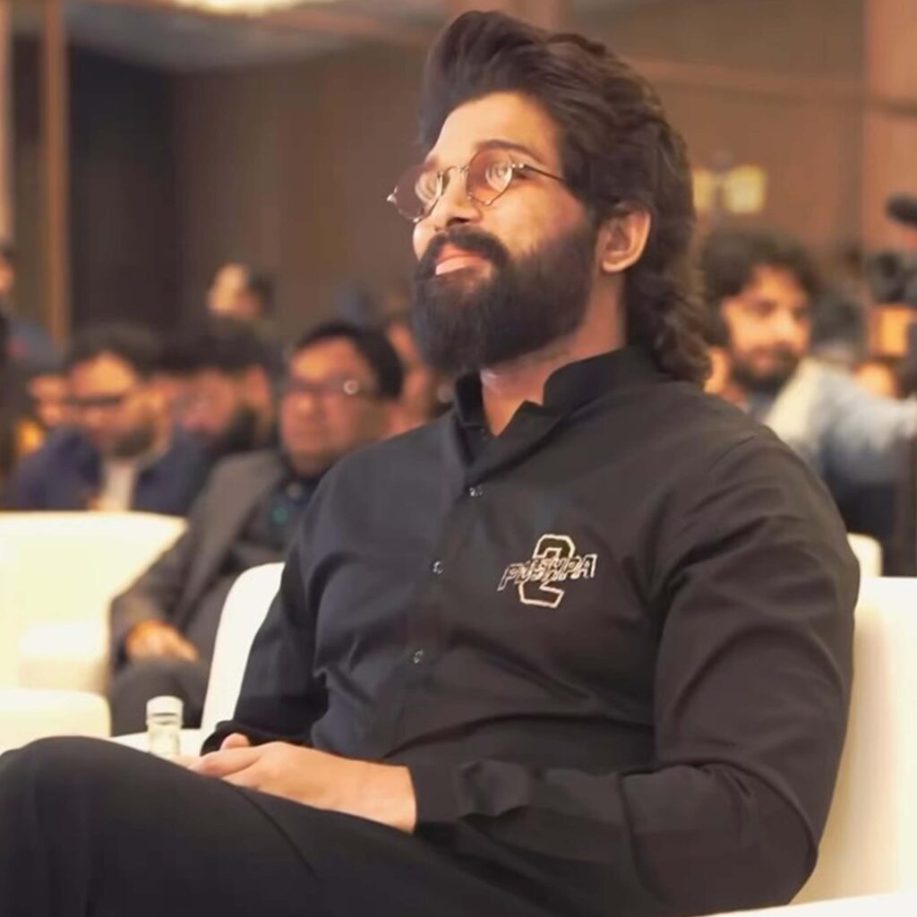 llu Arjun, dressed in a black shirt with the Pushpa 2 logo, sits attentively at an event with a confident expression and a stylish look, surrounded by a blurred audience in the background.